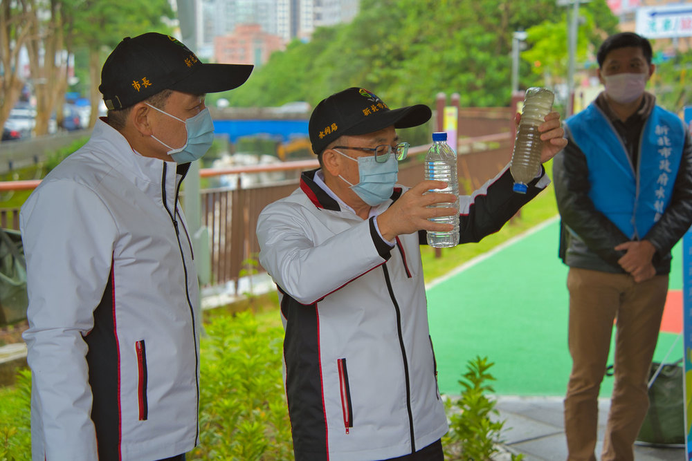 水利局長宋德仁(右)手持兩種寶特瓶，展示經過礫間處理前後的水質，市長侯友宜專心聆聽